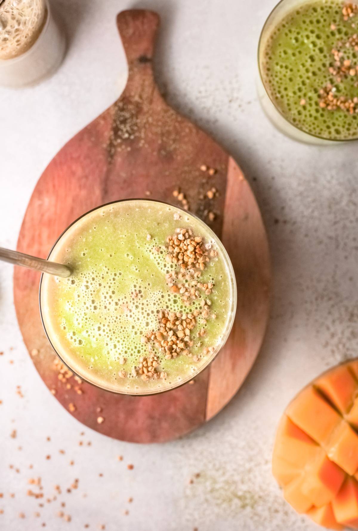 Green mango smoothie in a glass with a metal straw and sprinkled seeds, placed on a wooden board next to sliced melon—a refreshing vegan smoothie recipe.