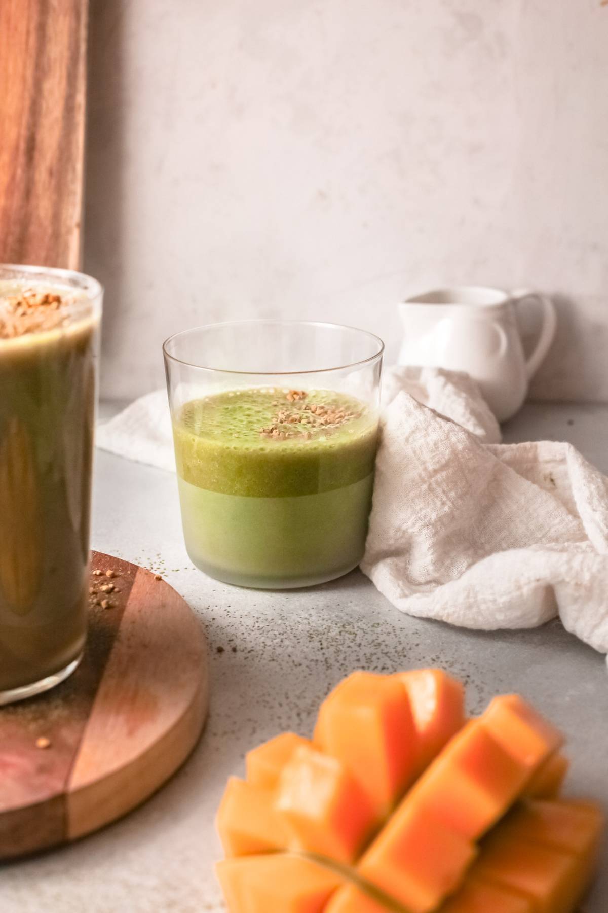 A glass of green mango smoothie sits on a table with a white napkin, sliced mango, and another drink nearby.
