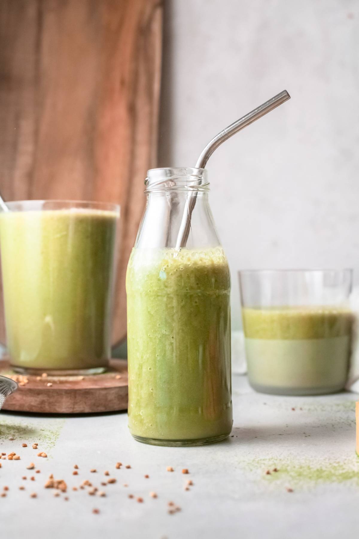 A glass bottle of green mango smoothie with a metal straw sits next to two glasses filled with vibrant green drinks, perfect for anyone looking for a refreshing vegan smoothie recipe.