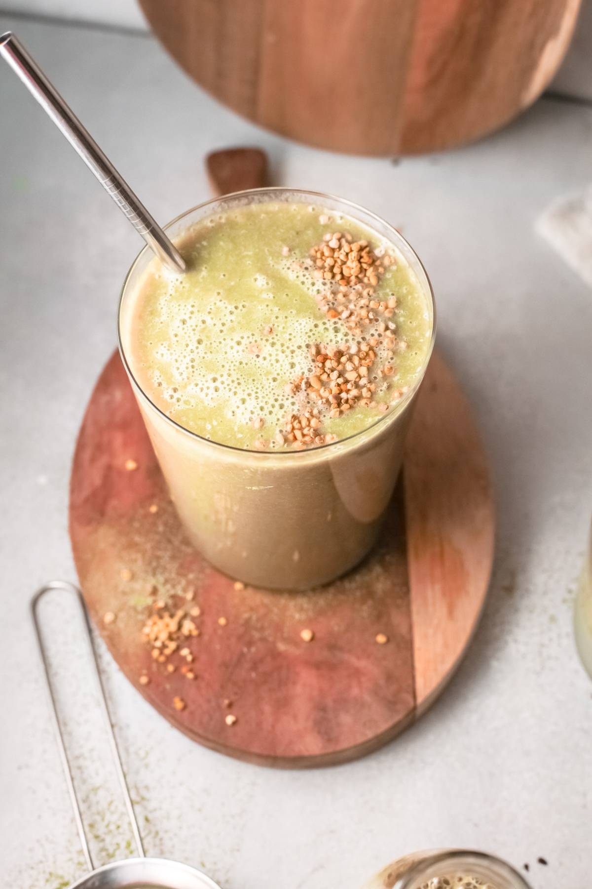 A Mango Matcha Smoothie in a glass with a metal straw, topped with sprinkled seeds, sits on a wooden board.