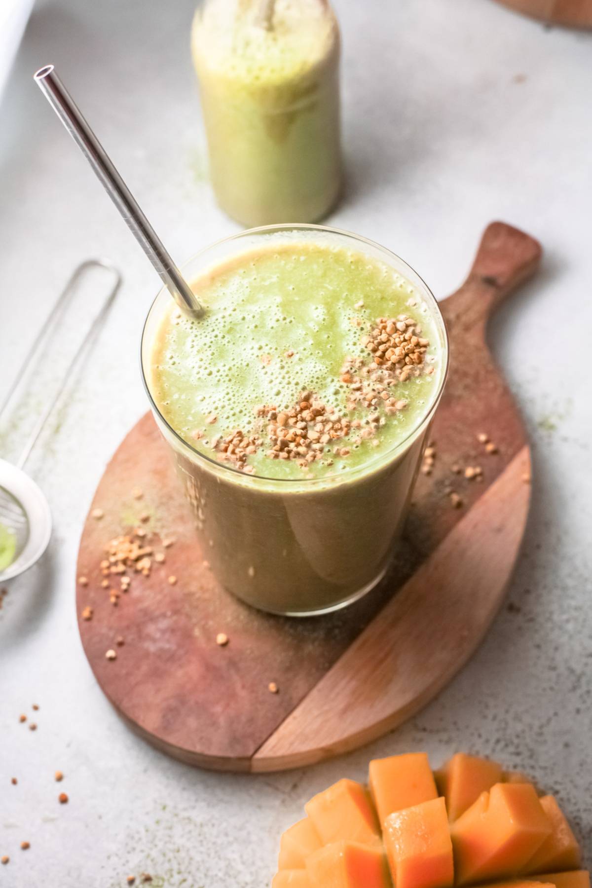 A glass of green mango smoothie with a metal straw and sprinkled toppings sits on a wooden board, beside cut fruit—a vibrant vegan smoothie recipe perfect for refreshing your day.