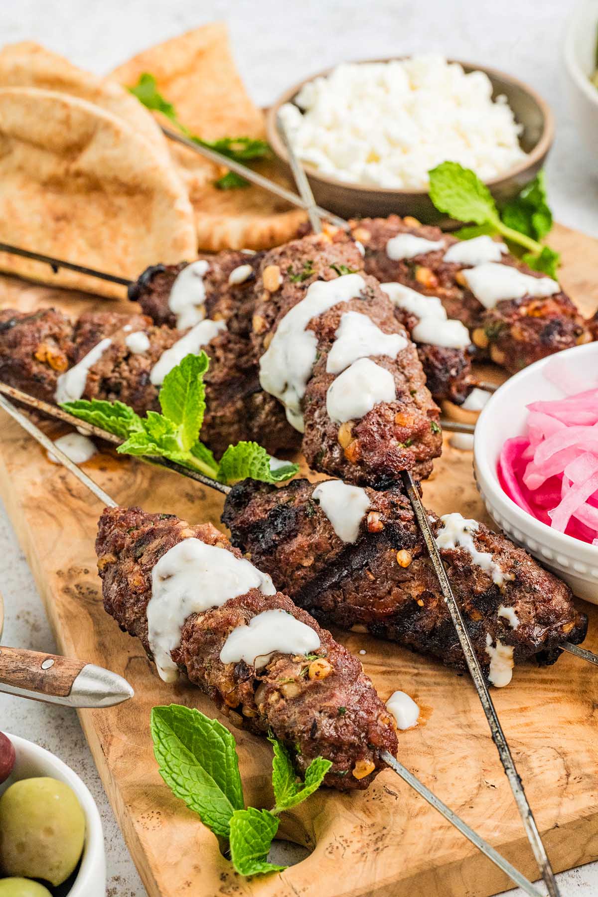 Grilled beef kofta kebabs on skewers with white sauce, fresh mint, pita bread, and pickled onions served on a wooden board.