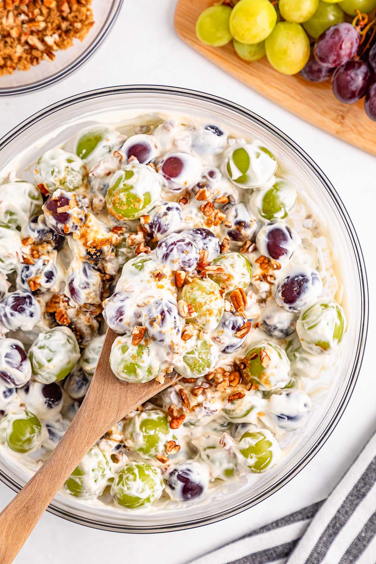 Bowl of creamy grape salad with green and red grapes, pecans, and a wooden spoon scooping out the delicious grape salad.