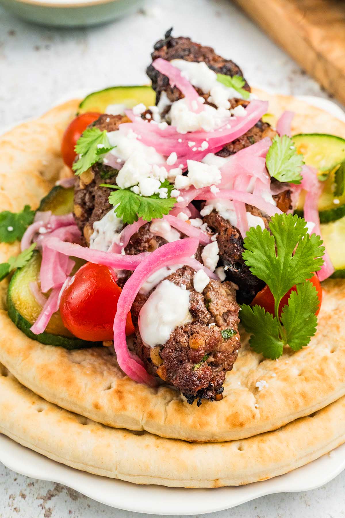 Pita bread topped with beef kofta kebabs, tomatoes, cucumbers, pickled onions, cilantro, feta, and white sauce.