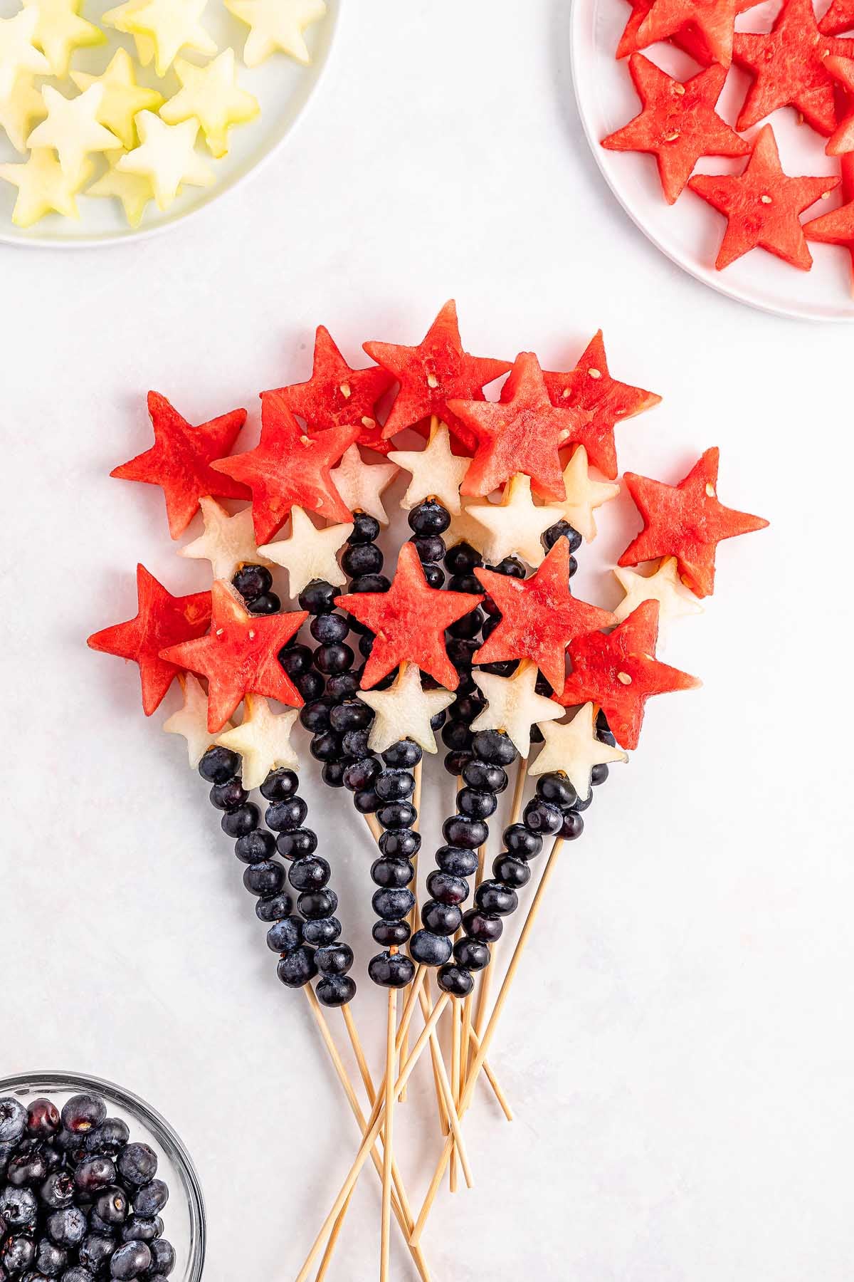 Watermelon Blueberry Sparklers feature fruit skewers with watermelon and melon star shapes on top, plus blueberries on sticks, all artfully arranged on a white surface.
