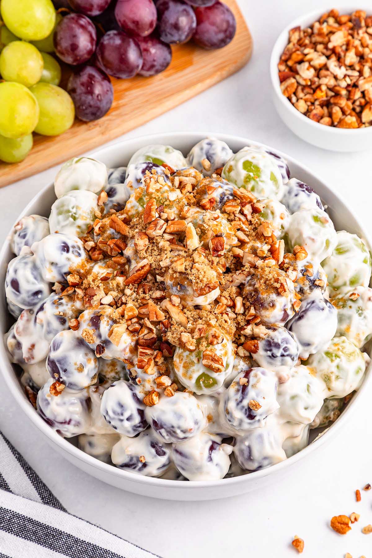 A bowl of creamy grape salad topped with chopped pecans, with fresh grapes and crunchy nuts in the background.