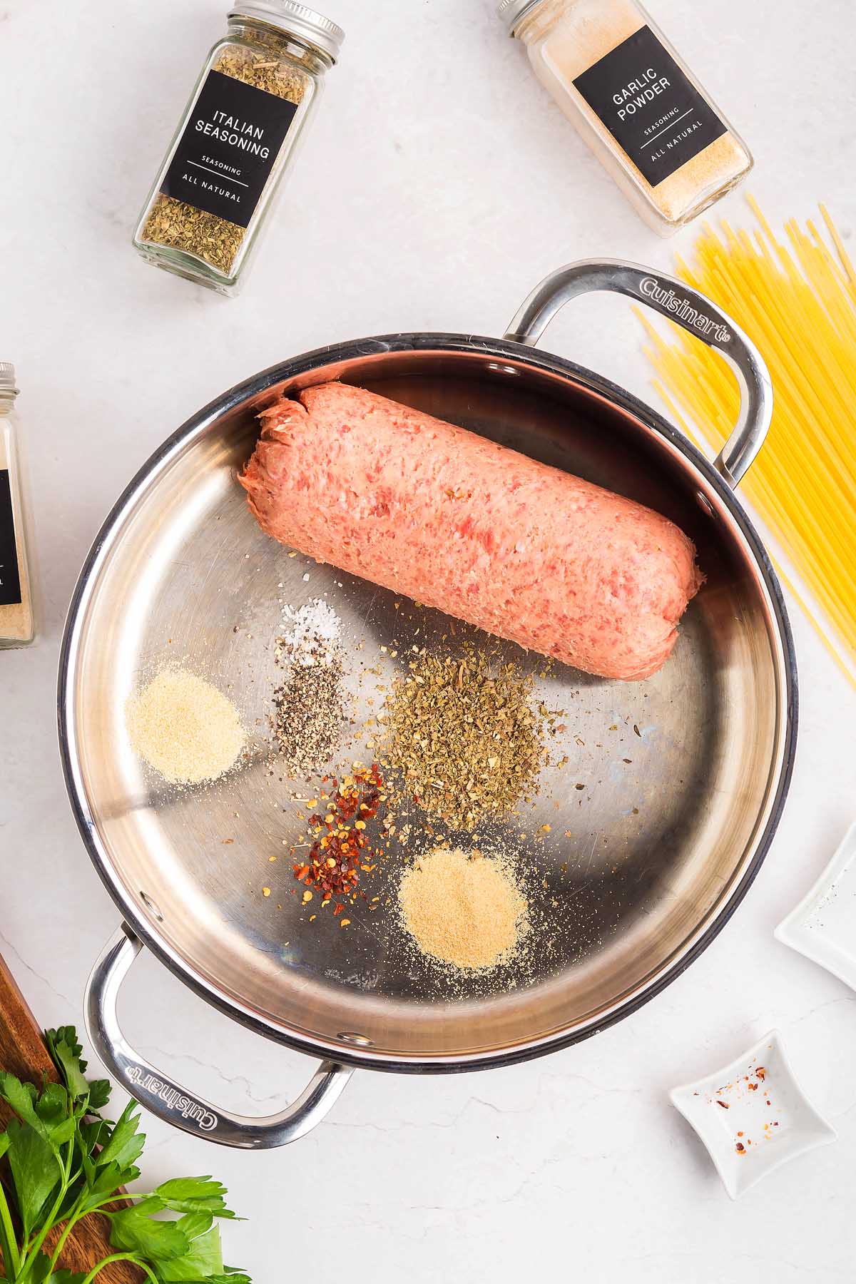 Raw ground meat and assorted spices in a metal pan, with spaghetti, seasoning jars, and hints of spaghetti pizza inspiration on a white surface.
