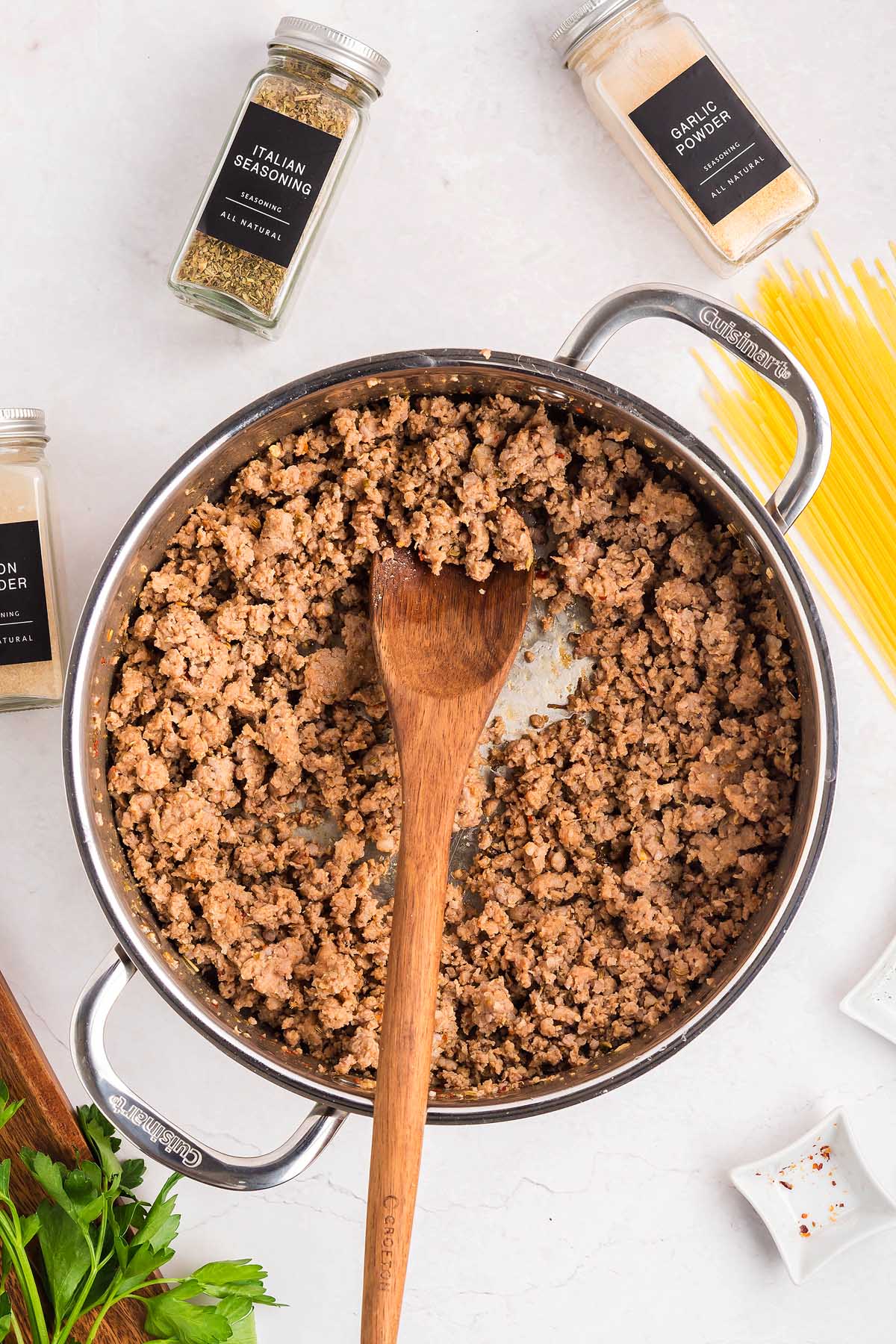 Cooked ground beef in a pot with a wooden spoon, surrounded by seasonings and uncooked spaghetti—perfect for crafting a delicious spaghetti pizza twist.