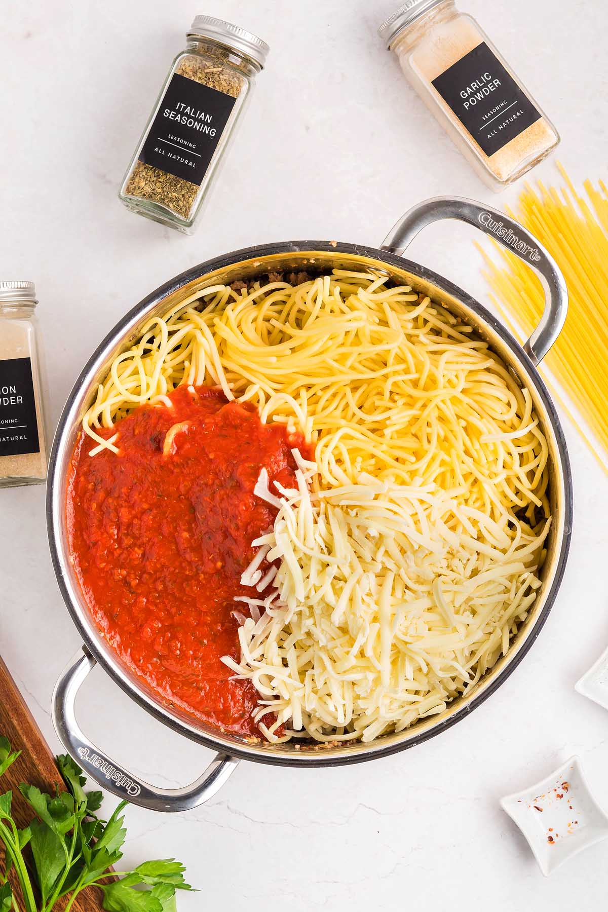 Pot of cooked spaghetti with tomato sauce and shredded cheese, surrounded by spice jars on a white surface—perfect for creating a spaghetti pizza-inspired meal.
