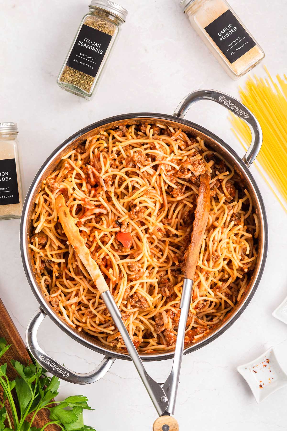 A pot of spaghetti with meat sauce sits on a white surface, surrounded by seasonings.
