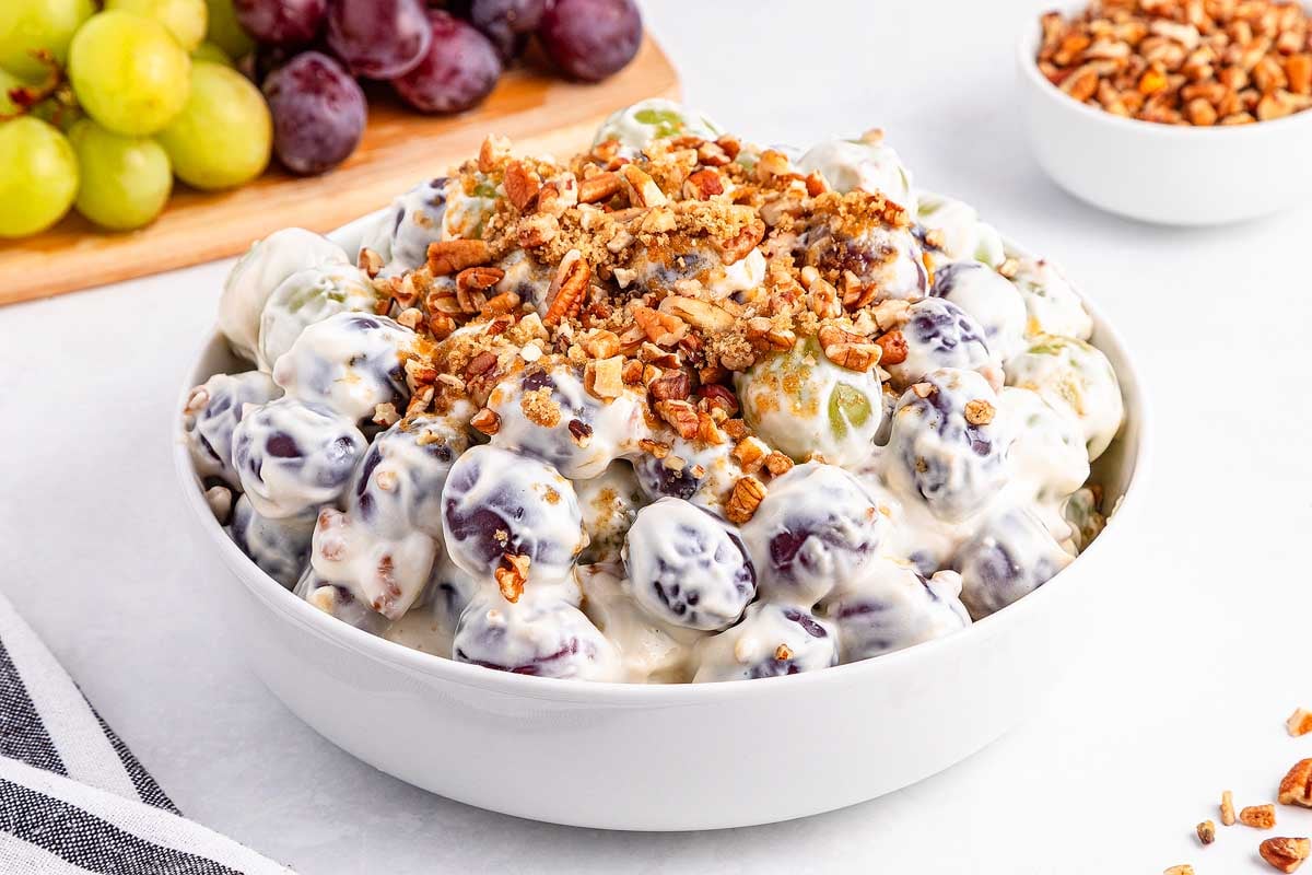 A bowl of grape salad topped with chopped pecans, with fresh grapes and nuts in the background.