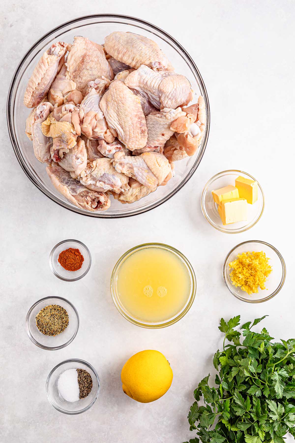 Raw chicken wings in a bowl, with small bowls of seasonings, butter, lemon, and fresh parsley on a white surface—perfect for making flavorful slow cooker lemon pepper wings.