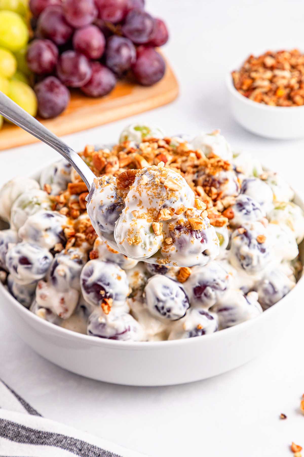 A bowl of creamy grape salad topped with chopped nuts, with a spoonful of the luscious grape salad being lifted out.