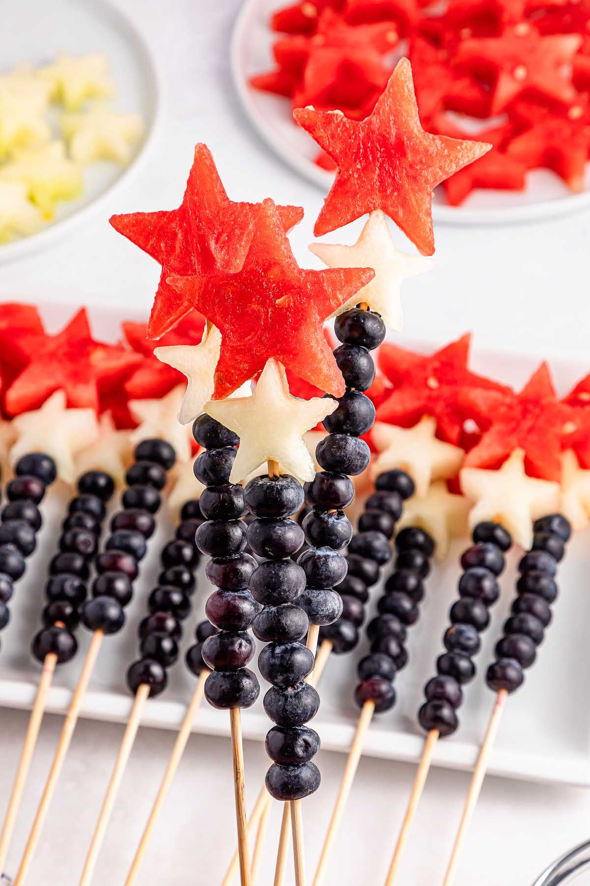 Watermelon Blueberry Sparklers: Fruit skewers featuring blueberries and star-shaped pieces of watermelon and melon arranged on a white surface.