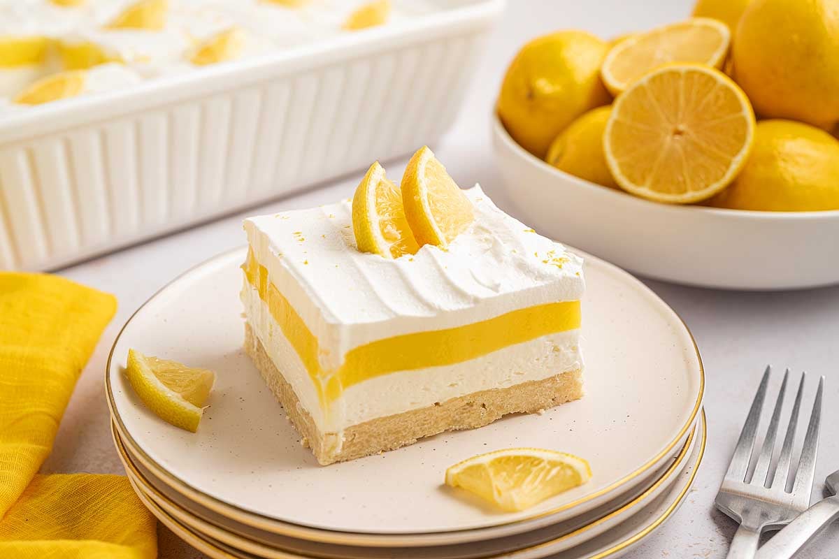 A lemon lush dessert bar with whipped topping and lemon slices, plated alongside fresh lemons and forks for a bright, zesty finish.