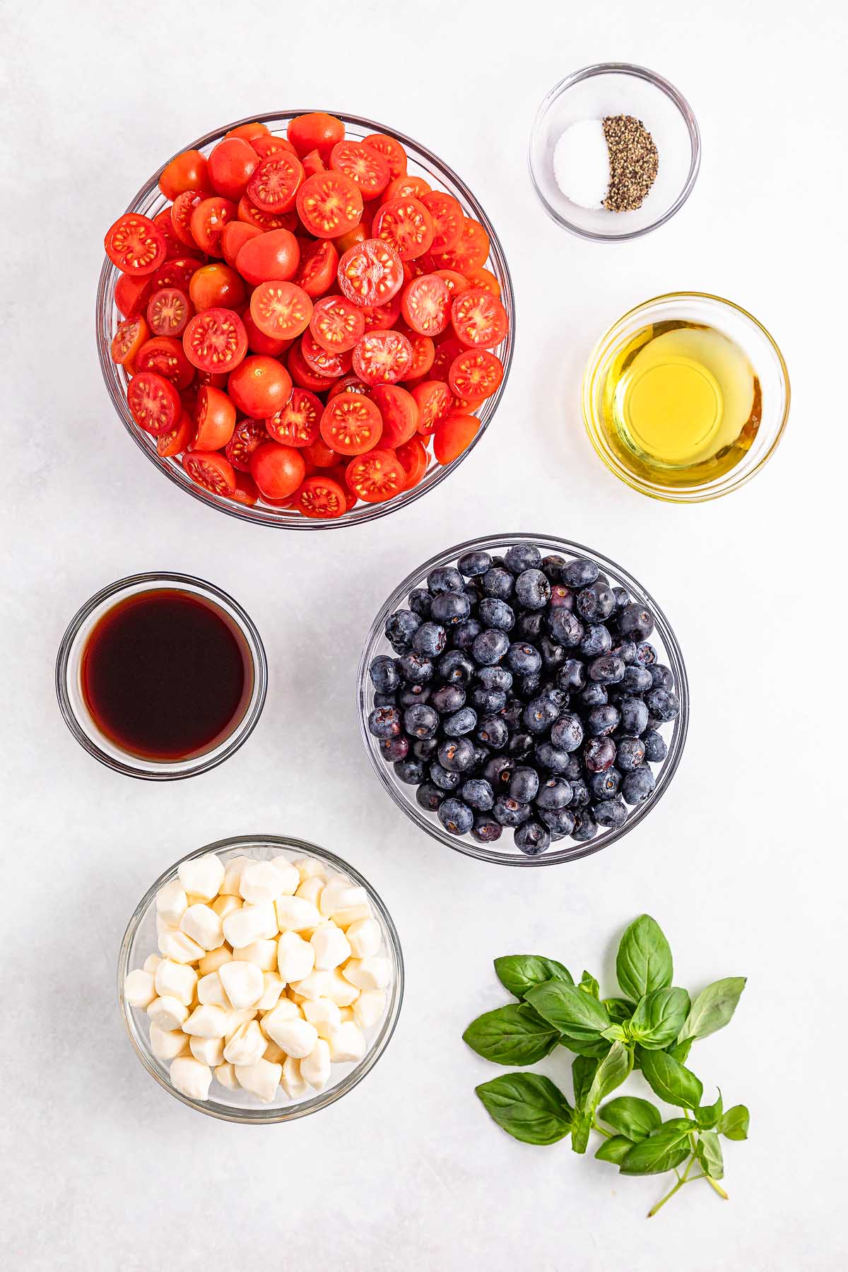 Bowls of cherry tomatoes, blueberries, mozzarella, vinegar, oil, salt, pepper, and fresh basil on a white surface create the perfect blueberry tomato Caprese.