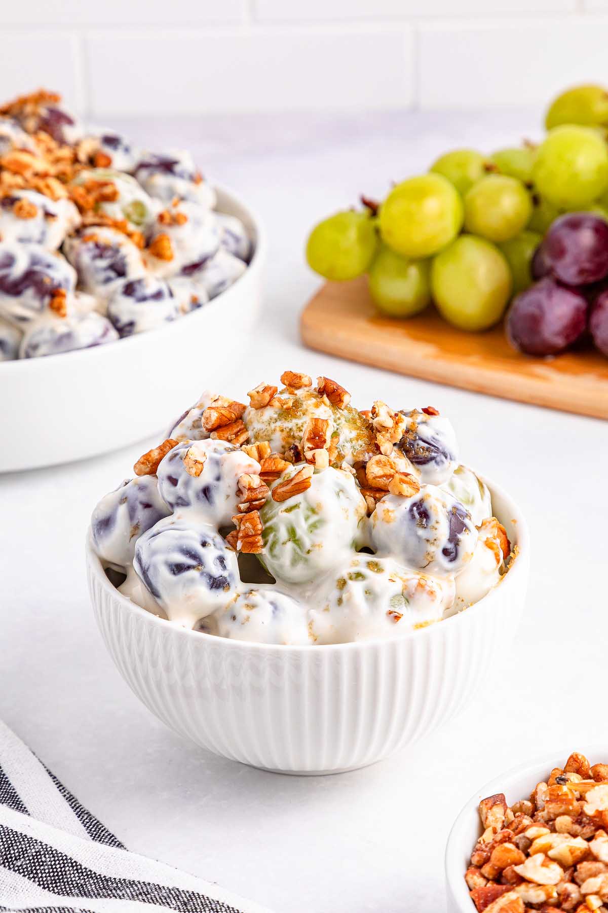 A bowl of creamy grape salad topped with chopped pecans, with fresh grapes and another bowl of grape salad in the background.