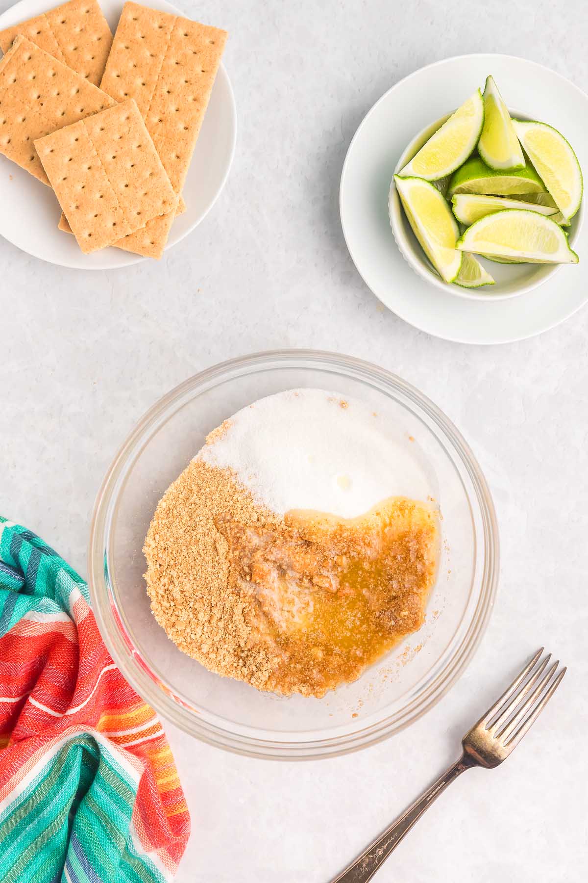 Glass bowl with graham crumbs, sugar, and butter beside graham crackers, lime wedges, and a fork—everything you need to start making delicious key lime bars. A striped towel adds a pop of color to the scene.