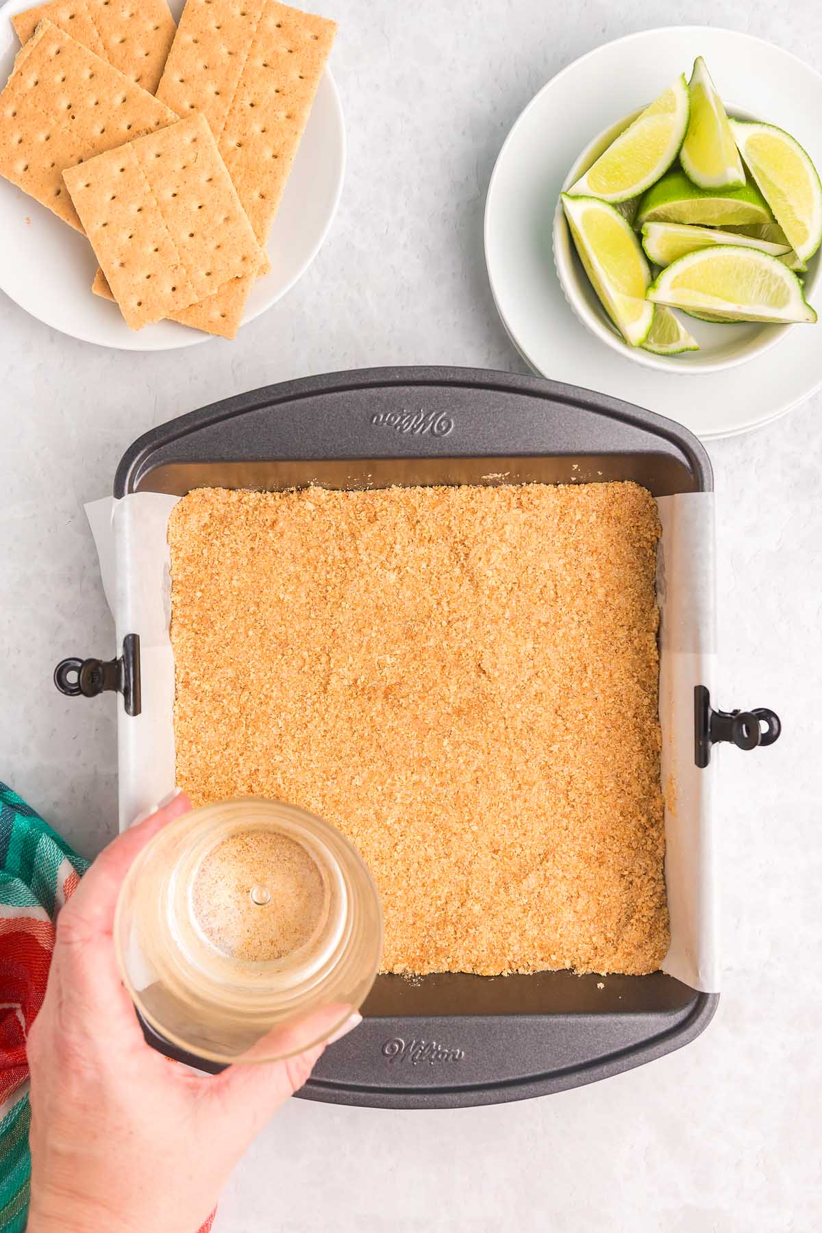Hand pressing graham cracker crust into a pan for key lime bars, with lime wedges and graham crackers on plates nearby.