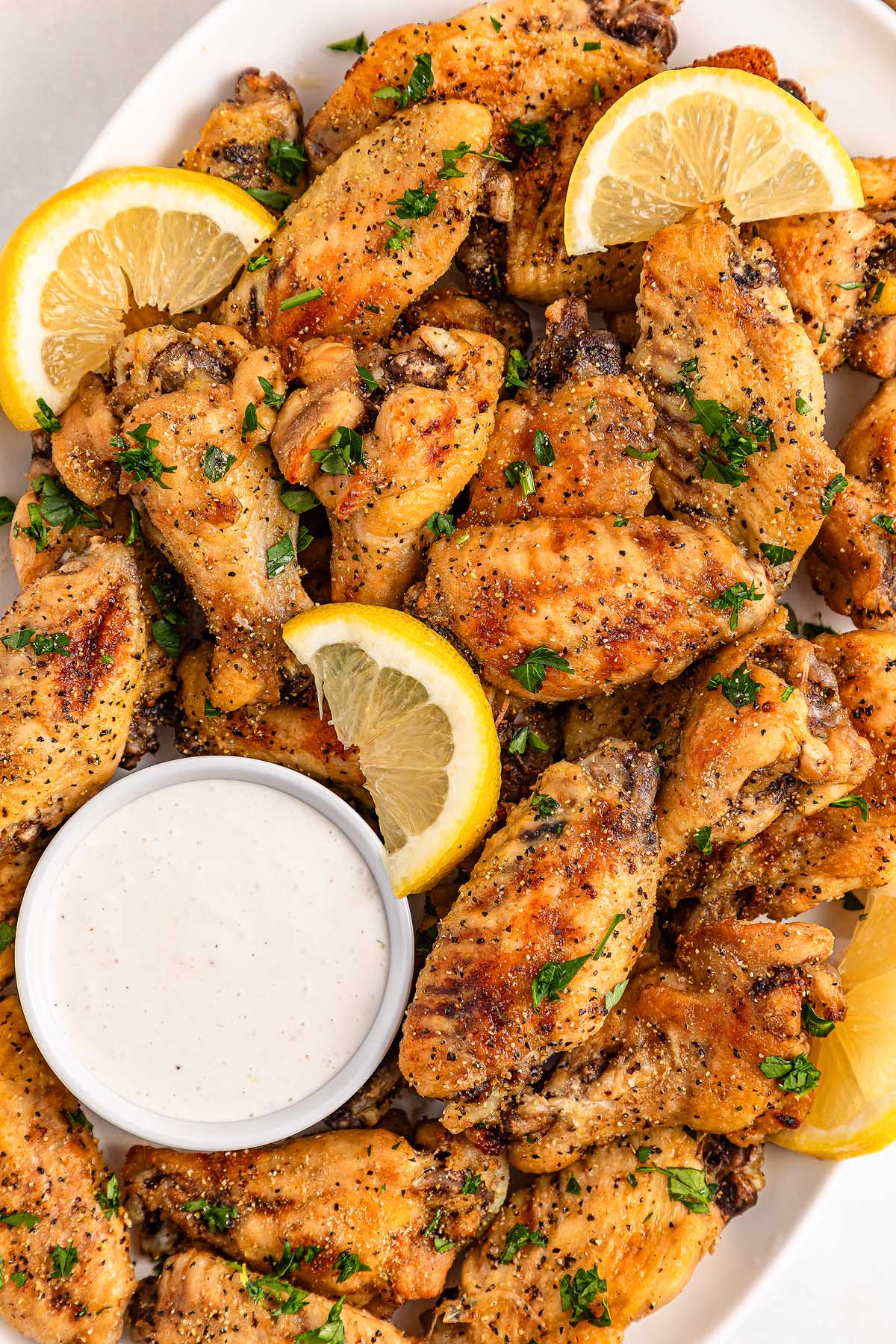 A plate of slow cooker lemon pepper wings with lemon slices, garnished with herbs, and served alongside a cup of ranch dip.