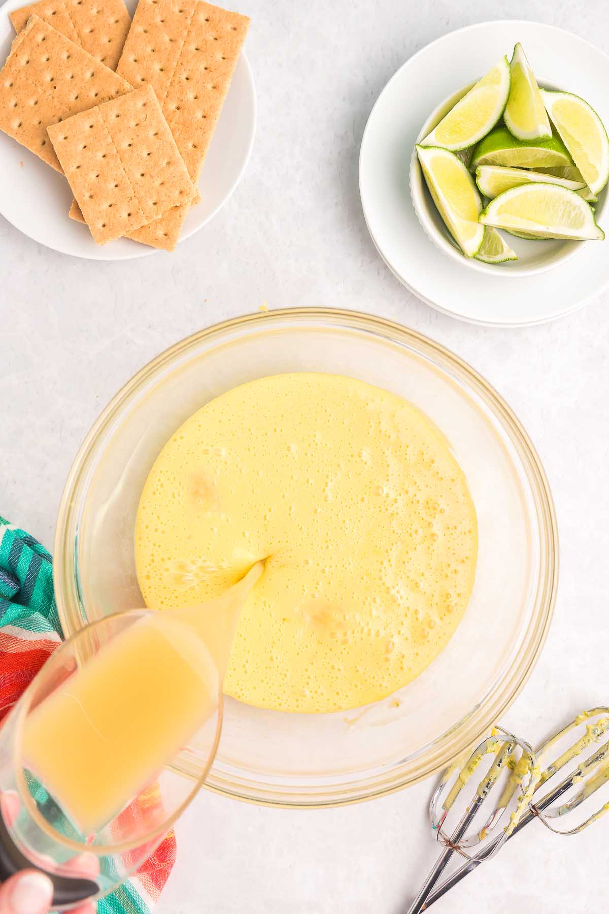 A hand pours juice into a bowl of yellow mixture, preparing key lime bars, with graham crackers and lime wedges nearby.