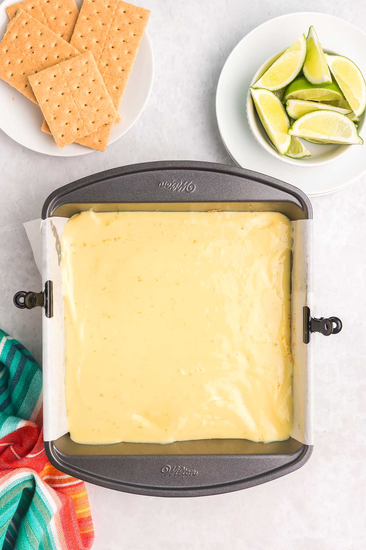Square pan with creamy yellow key lime bars batter, graham crackers, and lime wedges on plates beside it.