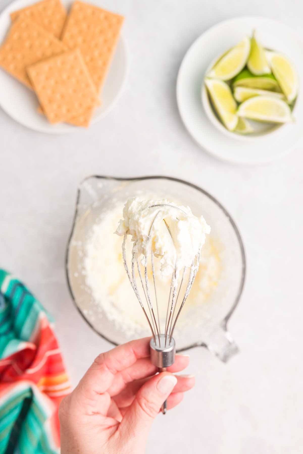 Hand holding a whisk with whipped cream, with graham crackers and lime wedges in the background—perfect for topping your freshly baked key lime bars.