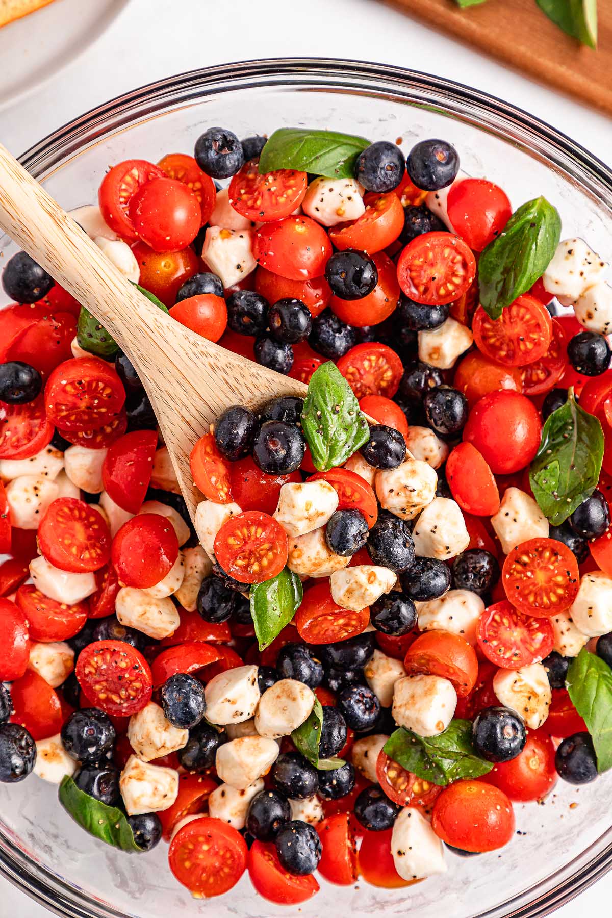 Blueberry tomato Caprese salad with mozzarella balls, basil, and a wooden spoon in a glass bowl.