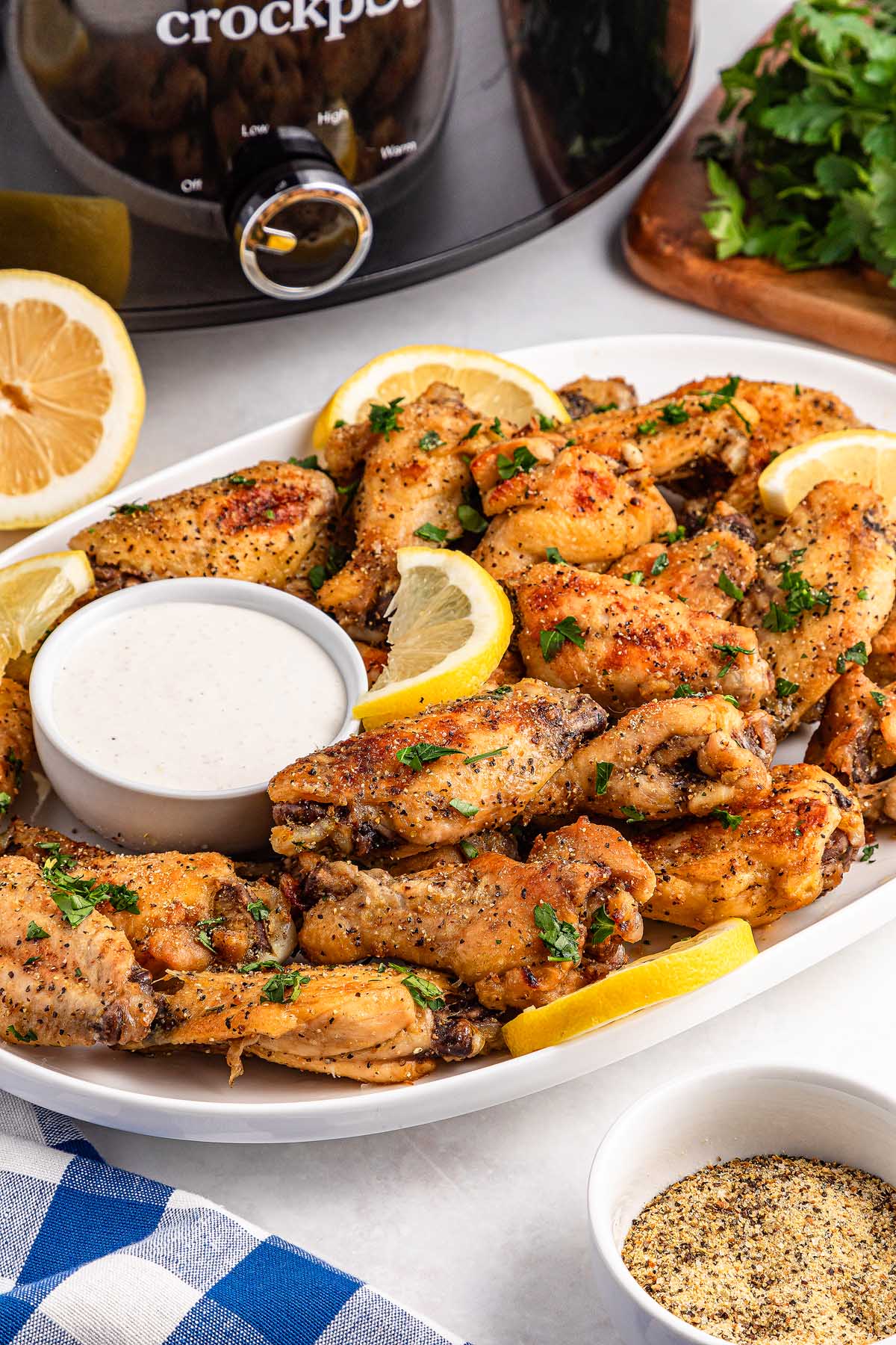 A plate of slow cooker lemon pepper wings is served with lemon slices and dipping sauce, with a crockpot in the background.