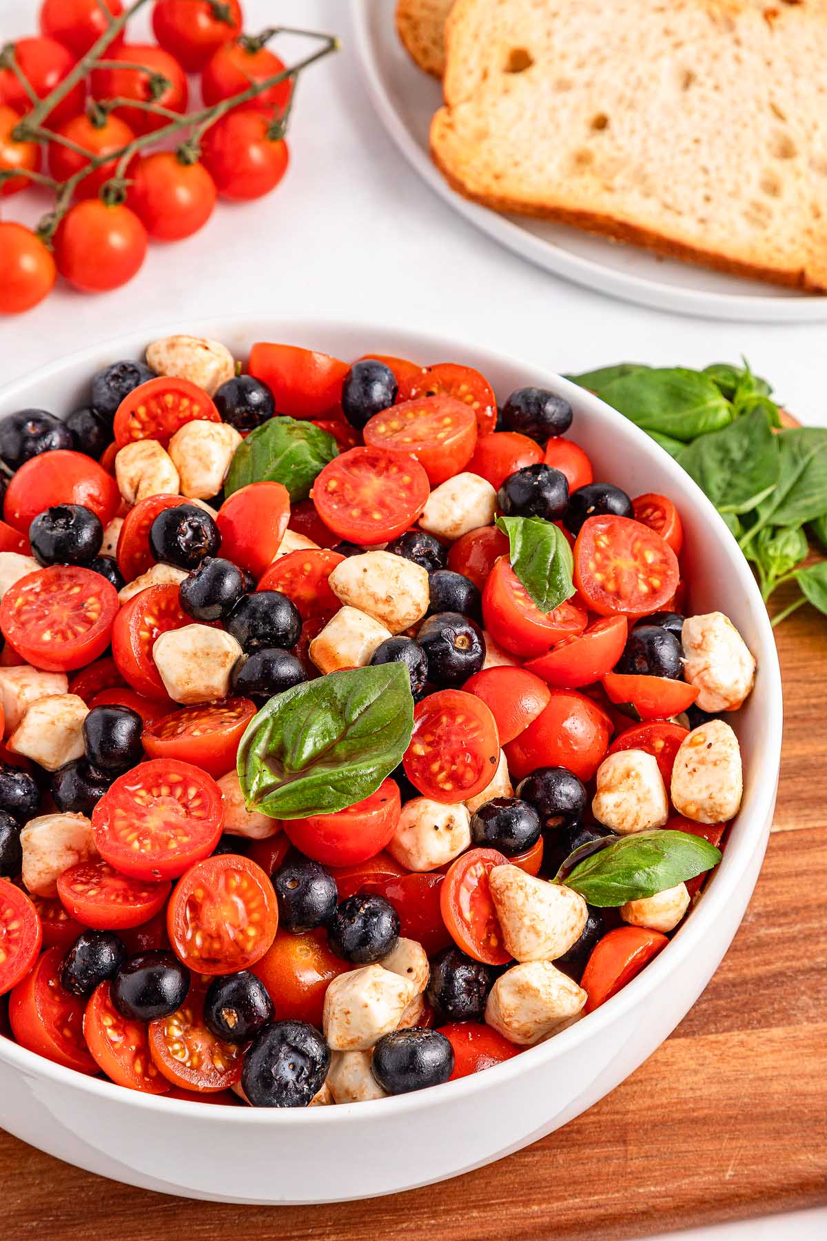 A bowl of Caprese salad with cherry tomatoes, blueberry, black olives, mozzarella balls, and fresh basil leaves.