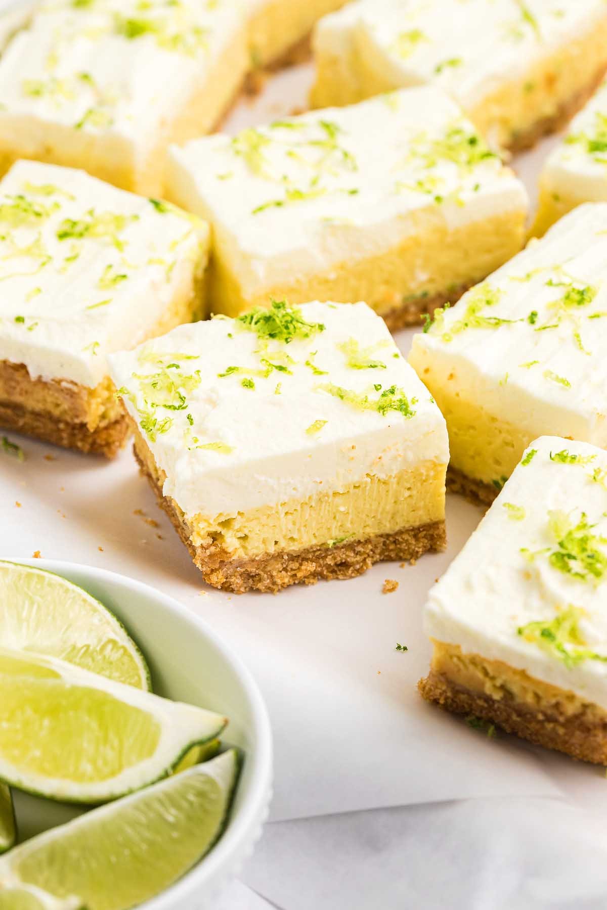 Key lime bars with a graham cracker crust, topped with fresh lime zest, are served next to a bowl of lime wedges.