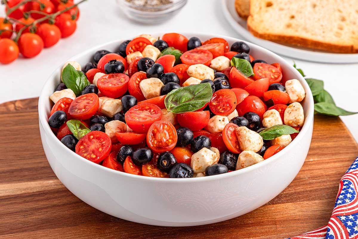 A bowl of blueberry tomato Caprese salad with black olives, mozzarella balls, and basil on a wooden board.