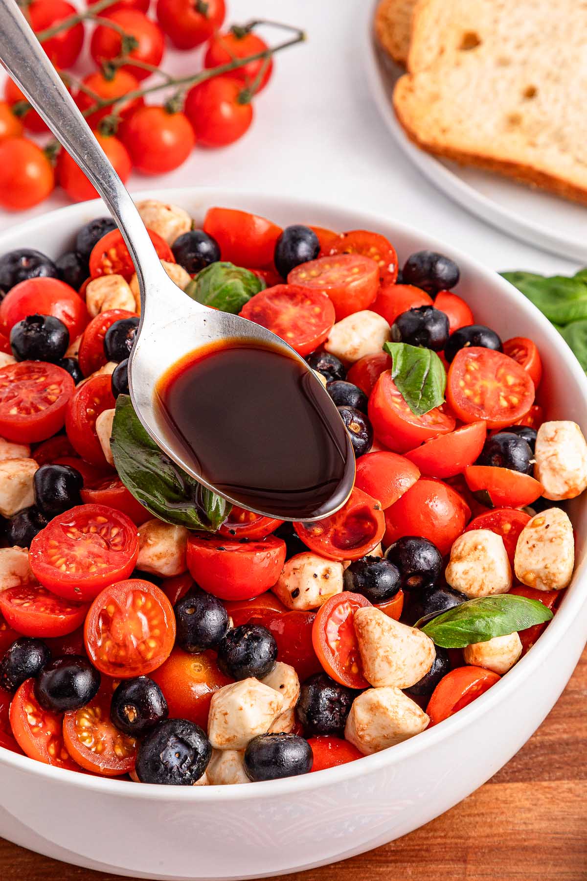 A spoon drizzles balsamic vinegar over a bowl of blueberry tomato Caprese with mozzarella and olives.
