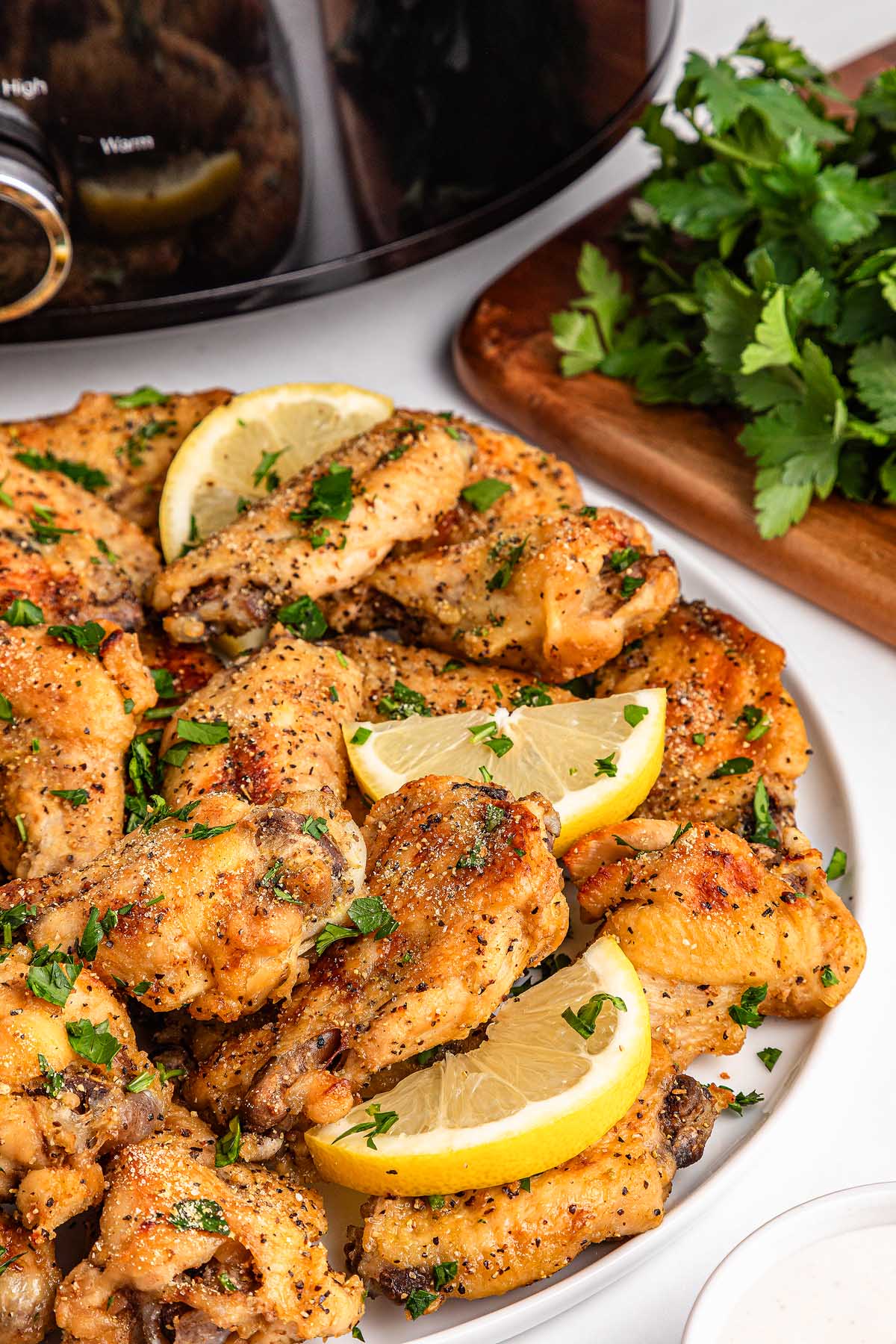 Plate of slow cooker lemon pepper wings, garnished with lemon wedges and chopped herbs, with extra fresh herbs and a slow cooker nearby.