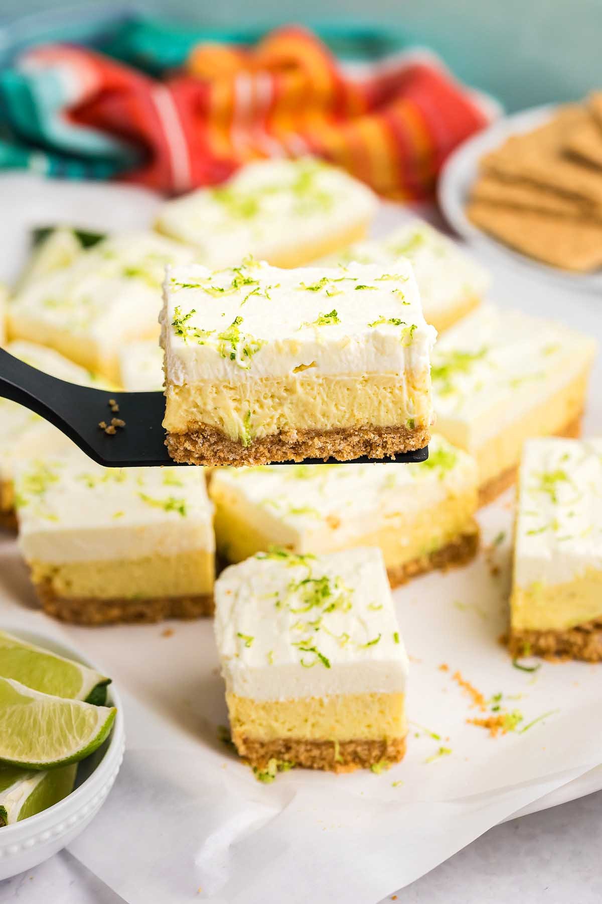 A spatula holds a creamy key lime cheesecake bar above more key lime bars and fresh lime wedges.