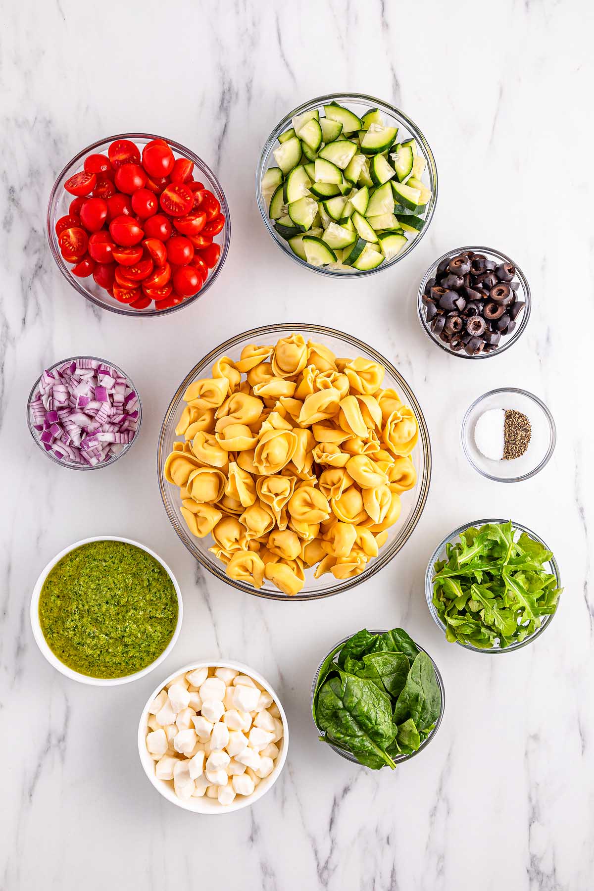 Bowls of pesto tortellini pasta salad with cherry tomatoes, zucchini, olives, red onion, spinach, mozzarella, and pepper on marble.