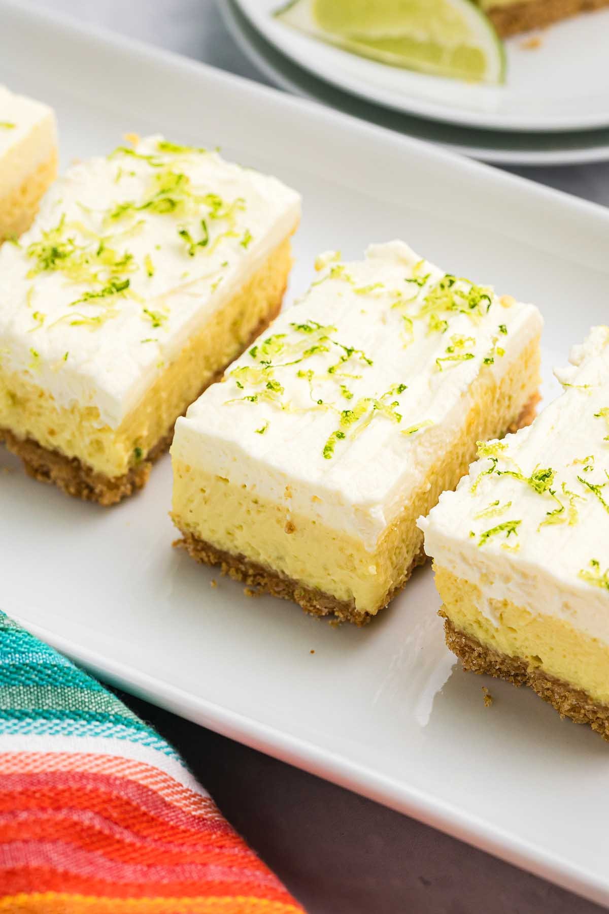 Key lime bars topped with whipped topping and lime zest sit on a white platter, next to a colorful striped napkin.