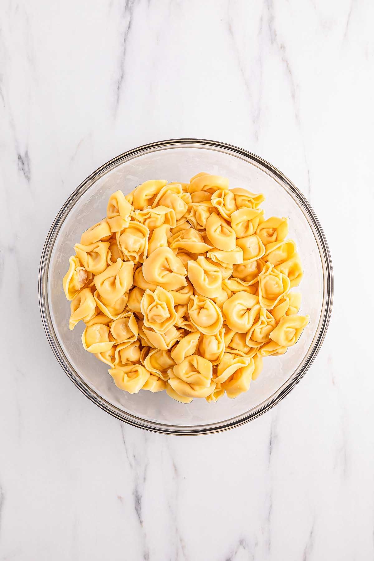 A glass bowl filled with uncooked cheese tortellini, perfect for making a fresh pesto tortellini pasta salad, sits on a white marble surface.