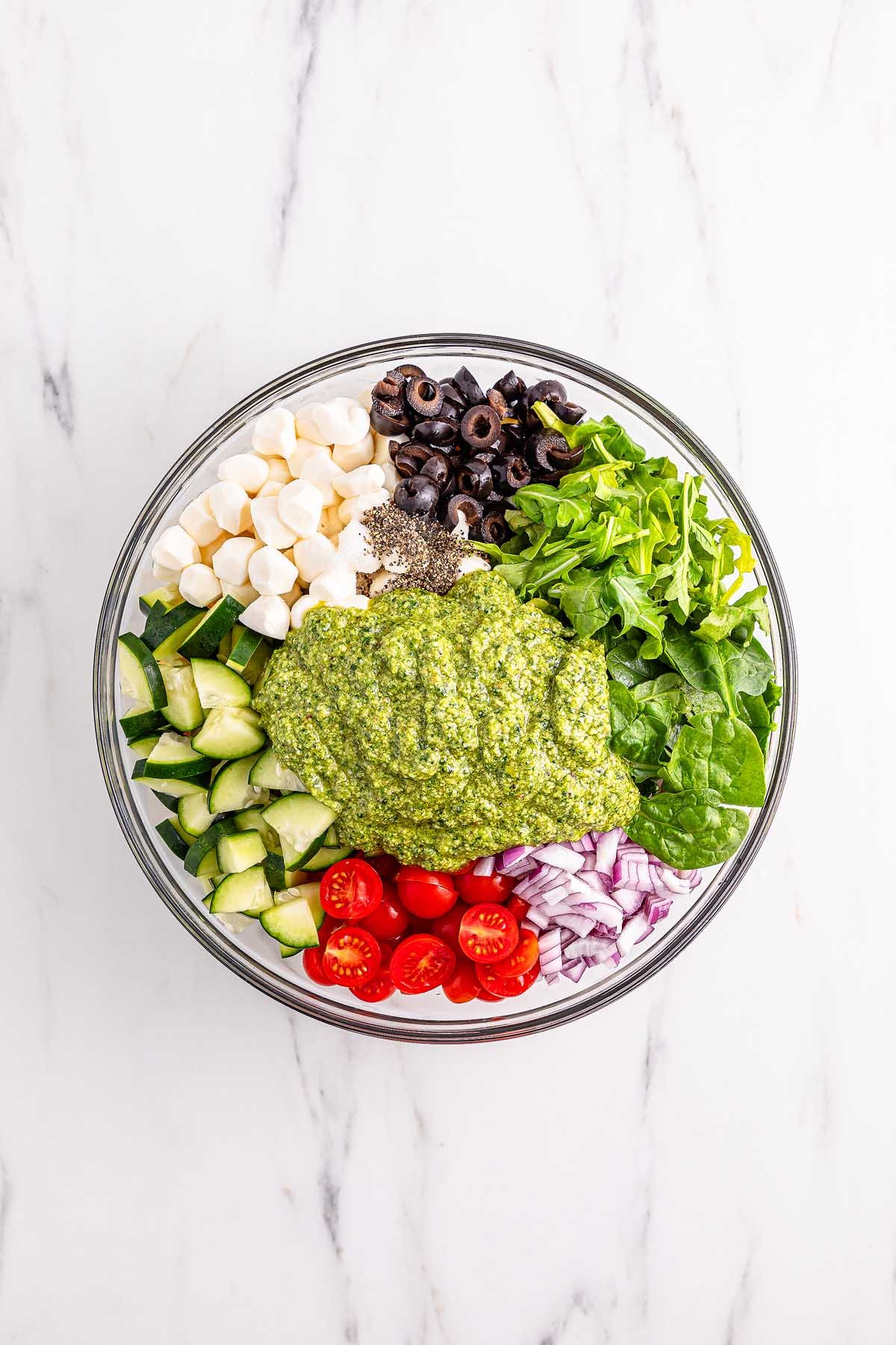 A glass bowl with pesto tortellini pasta salad, mozzarella, olives, arugula, spinach, cucumber, tomatoes, and red onion on a marble surface.