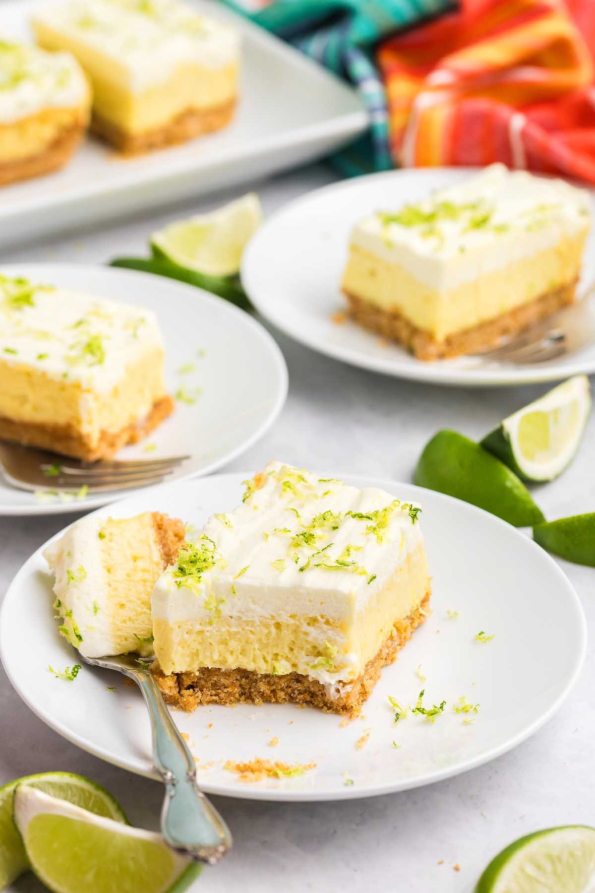 Three plates with creamy key lime bars, topped with whipped cream and lime zest, with limes nearby.