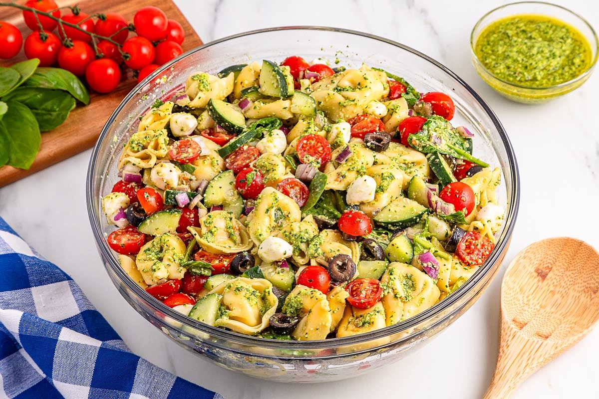 A bowl of pesto tortellini pasta salad with tomatoes, cucumber, olives, and mozzarella sits on a marble surface.