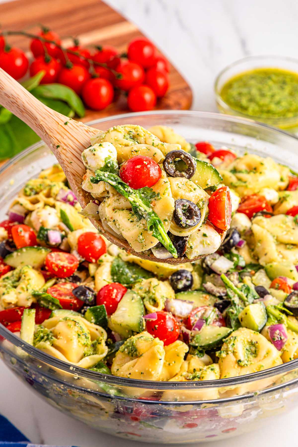 A wooden spoon holds pesto tortellini pasta salad with tomatoes, olives, and spinach above a glass bowl.