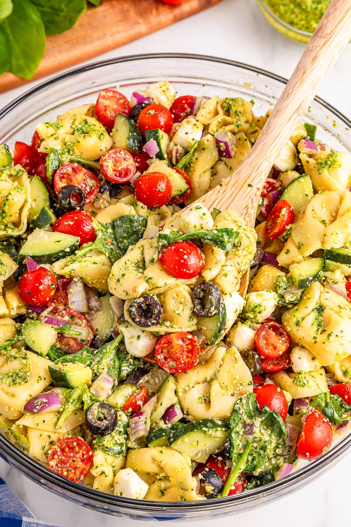 Bowl of pesto tortellini pasta salad with cherry tomatoes, spinach, olives, and a wooden spoon.
