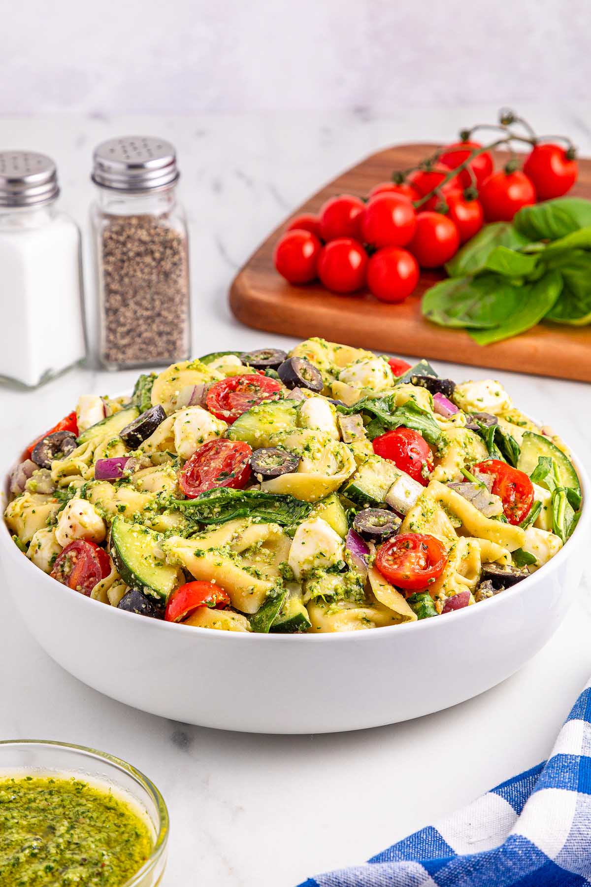 Bowl of pesto tortellini pasta salad with tomatoes, olives, mozzarella, and spinach on a white table surrounded by fresh ingredients.