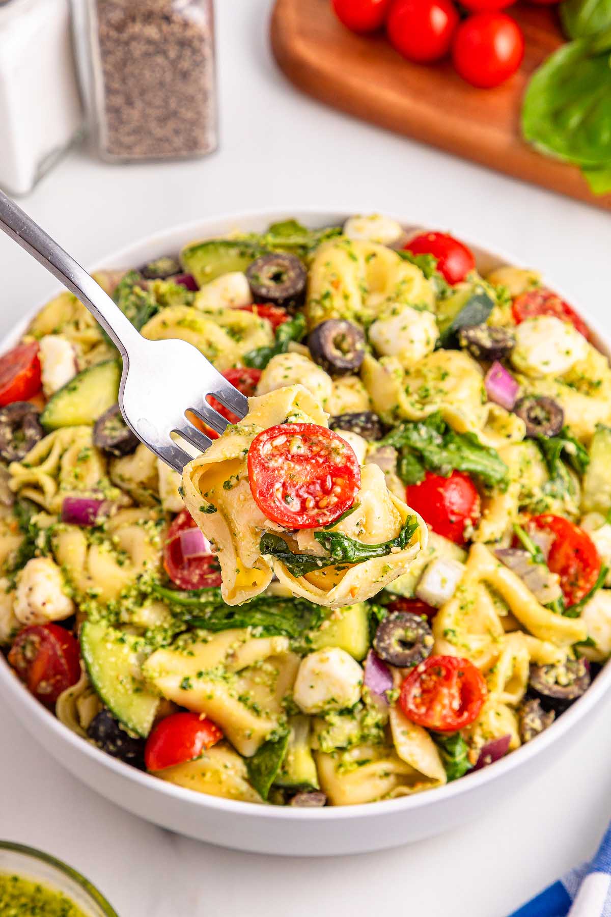 A fork holds pesto tortellini pasta salad with tomatoes, mozzarella, olives, and spinach above a bowl of the same vibrant salad.