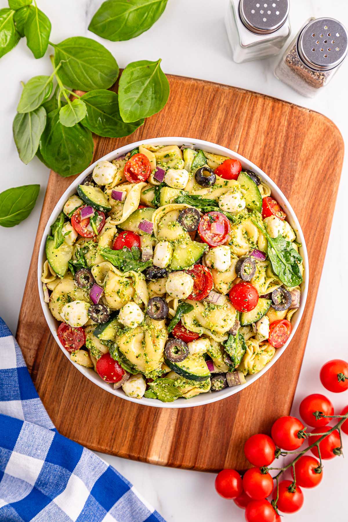 Bowl of pesto tortellini pasta salad with tomatoes, olives, mozzarella, and spinach on a wooden board.