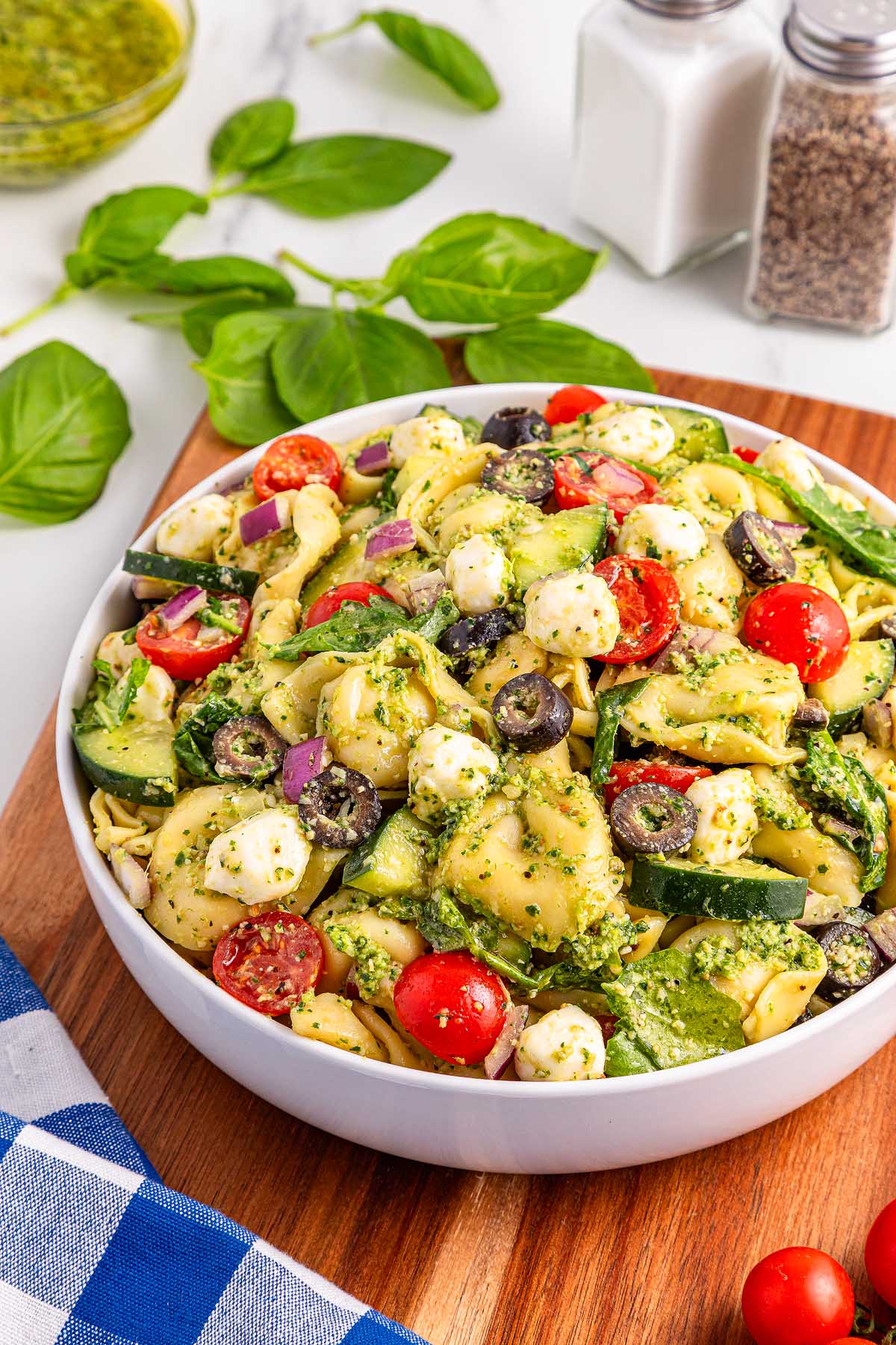 Bowl of pesto tortellini pasta salad with tomatoes, olives, mozzarella, and fresh herbs on a wooden board.