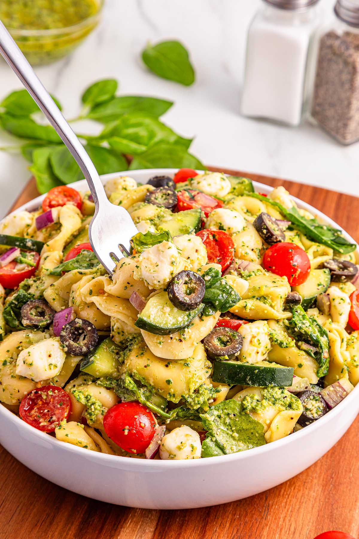 A fork lifts a serving of pesto tortellini pasta salad with tomatoes, olives, mozzarella, and fresh pesto in a white bowl.