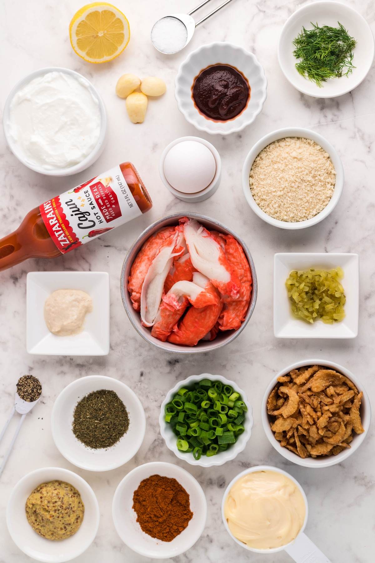 Various crab cake ingredients in bowls on a marble surface, including crab meat, spices, herbs, lemon, and the makings for a spicy sauce for crab cakes.