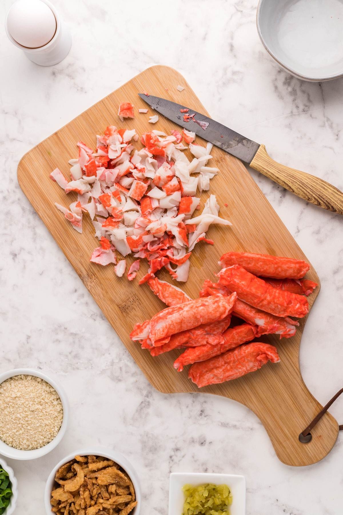 Chopped and whole imitation crab sticks on a wooden cutting board with a knife, surrounded by small bowls of ingredients—perfect for whipping up crab cakes in an air fryer or serving with a spicy sauce for crab cakes.